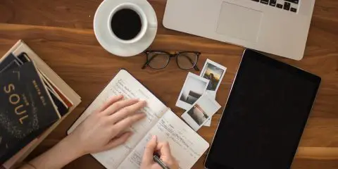 woman writing in a journal with a cup of coffee next to her on the table and a pair of glasses